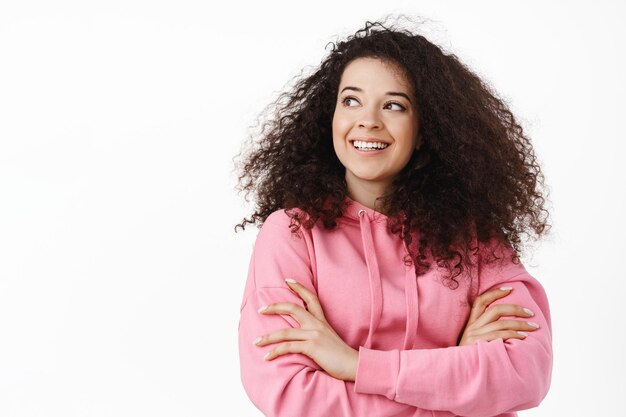 Close up portrait of stylish modern girl with curly hair, wearing pink hoodie, cross arms on chest, smiling and looking aside at logo, standing over white background.