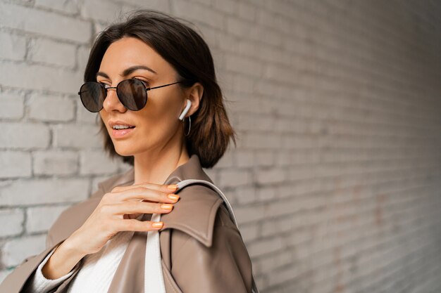 Close up portrait of Stylish short haired woman in casual leather coat and sunglasses posing over urban brick wall