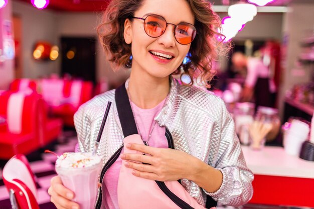Крупный план портрета стильной улыбающейся красивой женщины в ретро-винтажном кафе 50-х годов, пьющей коктейль с молочным коктейлем в розовой футболке и серебряной куртке в розовых солнцезащитных очках, веселящейся в веселом настроении