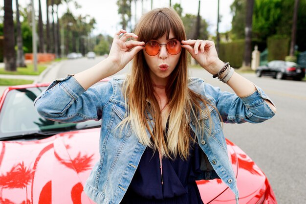 Close up portrait of surprised brunette woman sitting on the hood of amazing red convertible sport car in California.