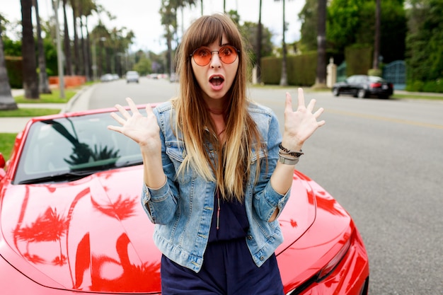 Close up portrait of surprised brunette woman sitting on the hood of amazing red convertible sport car in California.