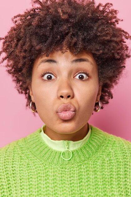 Close up portrait of surprised curly haired woman keeps lips folded wants to kiss you blows mwah at camera wears casual green knitted jumper isolated over pink background. Human face expressions