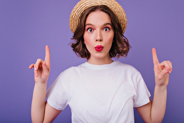 Close-up portrait of surprised dark-eyed girl in summer hat. Indoor shot of funny curly female model in white t-shirt posing with fingers up on purple wall.