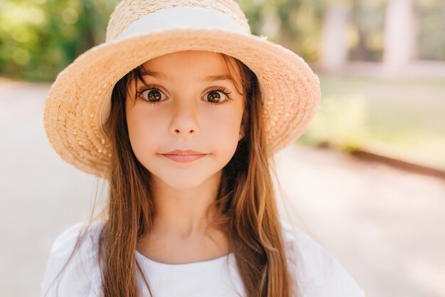 Close-up portrait of surprised kid with big shiny brown eyes posing. Amazing little girl in trendy summer hat standing on the road in sunny day.