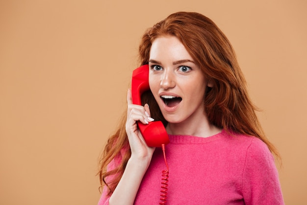 Close up portrait of a surprised pretty redhead girl talking by classic red phone