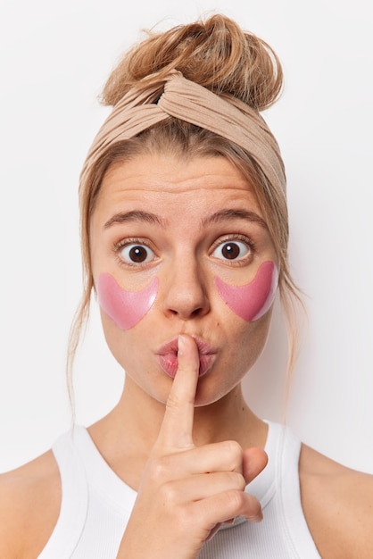 Close up portrait of surprised woman with shocked expression applies pink patches under eyes makes silence gesture tells secret of beauty wears beige headband isolated over white background.