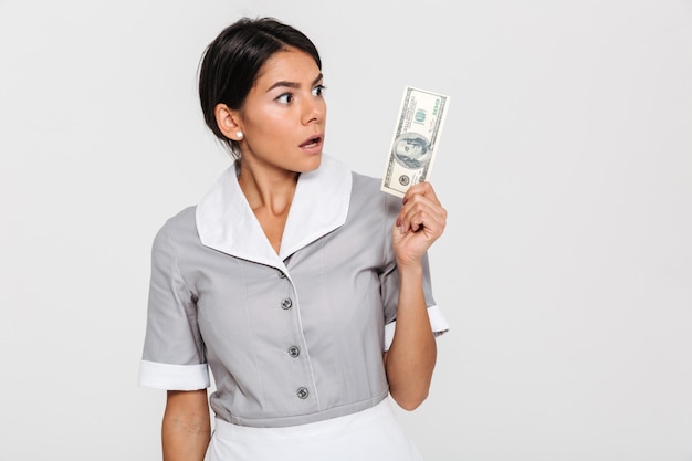 Foto gratuita ritratto del primo piano della giovane donna sorpresa in uniforme che tiene una banconota da cento dollari