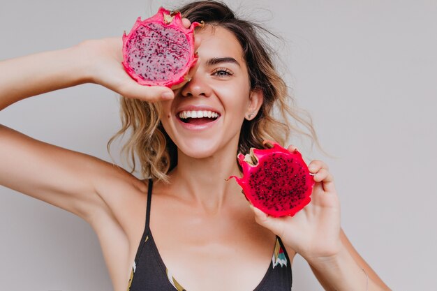 Close-up portrait of tanned girl with light eyes holding dragon fruit near face. Indoor shot of cute curly woman posing with tasty red pitaya.