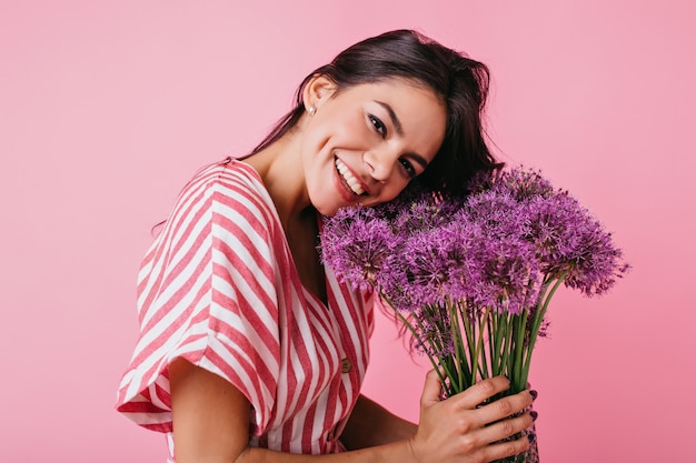 Close-up portrait of tanned woman with charming dimples on her cheeks. Girl is smiling cute, tilting her head to flowers.