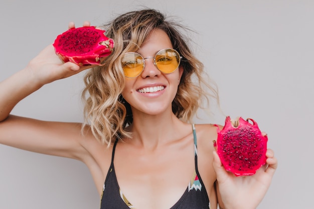 Close-up portrait of tanned woman in yellow sunglasses posing with red pitahaya. Indoor shot of happy caucasian girl holding exotic fruits and smiling.