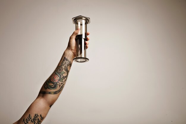 Close up portrait of a tattooed man's hand holding clear light gray aeropress nontraditional coffee brewing equipment high in the air isolated on white