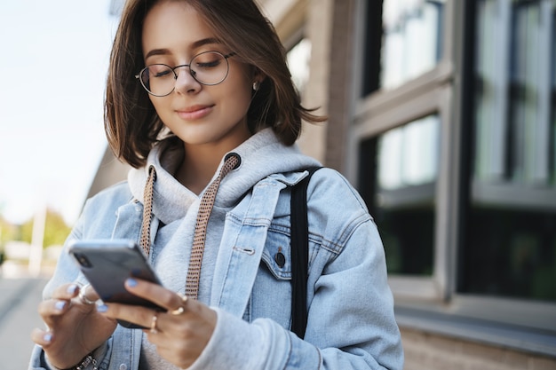 Close-up portrait tender romantic girlfriend send cheerful message, heart emoji friend, holding mobile phone and smiling at display, texting with friend, communicating while standing on street.