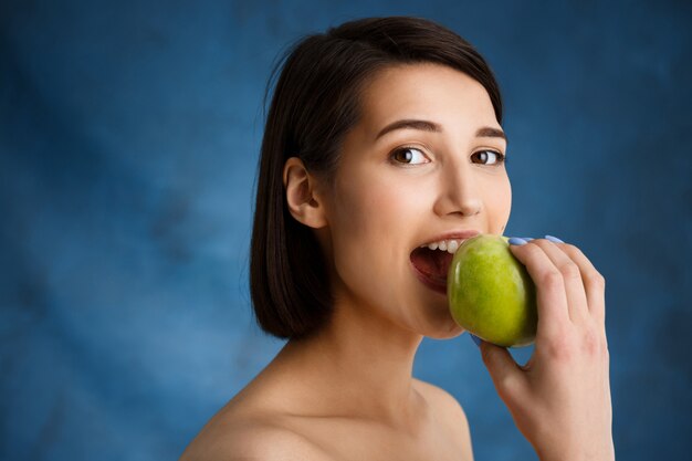 Close up portrait of tender  young woman biting apple over blue wall