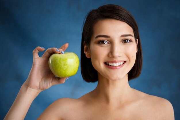 Close up portrait of tender  young woman holding apple over blue wall