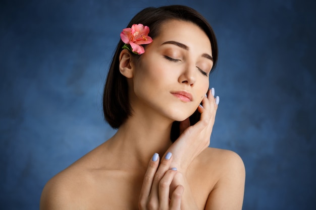 Foto gratuita chiuda sul ritratto di giovane donna tenera con il fiore rosa in capelli sopra la parete blu