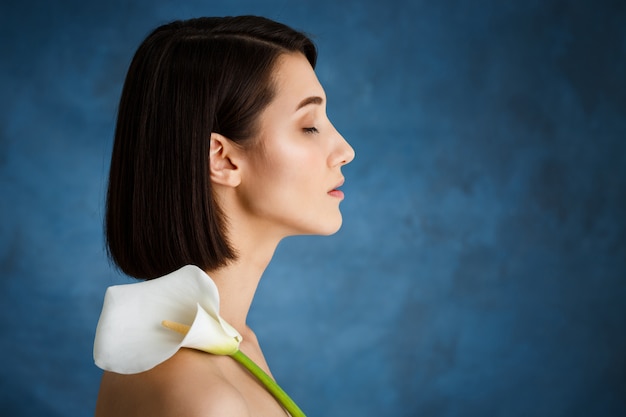 Chiuda sul ritratto di giovane donna tenera con il fiore bianco sopra la parete blu