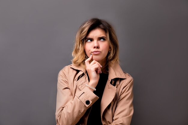 Close-up portrait of thoughtful look girl with big brown eyes looking away. Indoor photo of amazed european woman with short light hair isolated on grey wall.