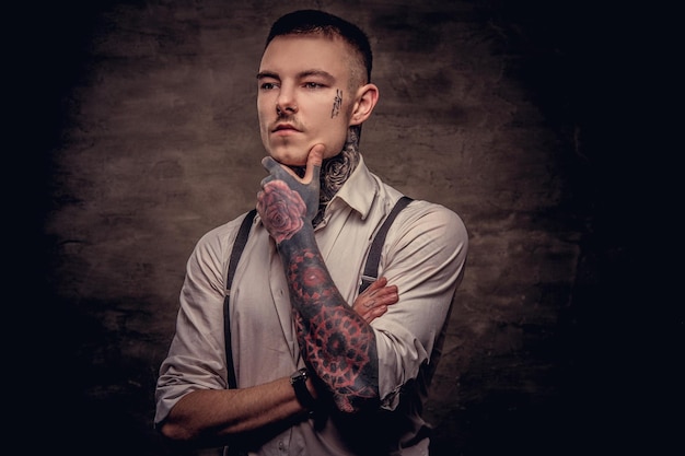 Free photo close-up portrait of a thoughtful young old-fashioned tattooed guy wearing white shirt and suspenders holds hand on chin. isolated on a dark background.