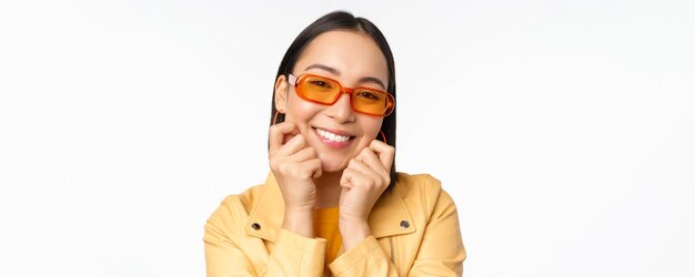 선글라스를 끼고 흰색 배경 위에 서 있는 카메라를 바라보며 낭만적인 미소를 짓고 있는 얼굴을 만지고 있는 트렌디한 아시아 여성의 초상화를 클로즈업