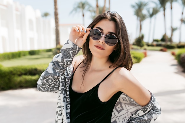 Free photo close-up portrait of trendy girl wearing sunglasses and stylish shirt, posing in the park and enjoying southern sun in vacation. amazing young woman with dark straight hair standing outside.