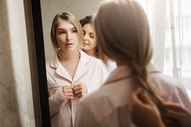 Free photo close-up portrait of two beautiful women at home. attractive young blonde standing near mirror, changing clothes from pyjamas and waiting while mother combing braid. typical cozy morning in family