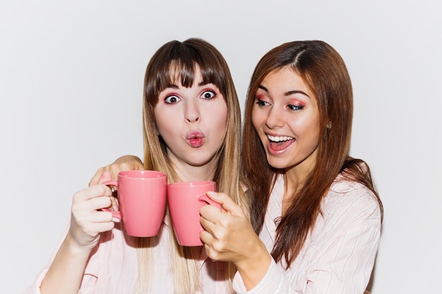 Close up portrait of Two cheerful white  women in pink pajamas with cup of tea posing. Flash portrait.