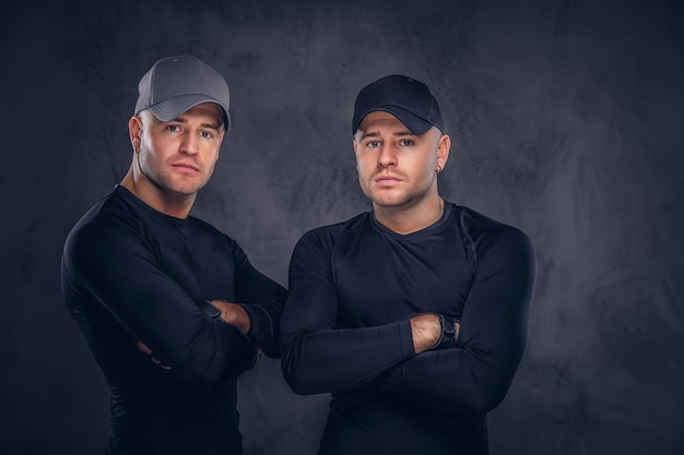 Close-up, a portrait of two handsome young male dressed in a black sportswear and baseball cap over a dark background.