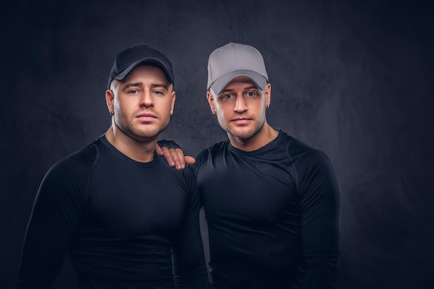 Close-up, a portrait of two handsome young male dressed in a black sportswear and baseball cap over a dark background.