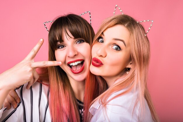 Close up  portrait of two happy exited woman wearing cat party hair accessories, bright make up, funny crazy emotions, friends enjoying party, pink wall