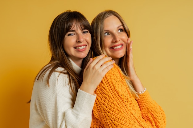 Close up portrait of two pretty woman in cozy sweaters posing on yellow. Autumn and winter fashion trends.