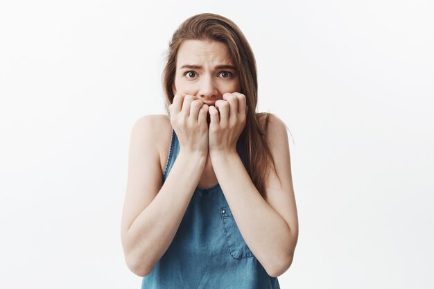 Close up portrait of unhappy young caucasian dark-haired srudent girl in blue shirt holding hands near face,  with cared expression. Woman being frightened watching scary movie with b