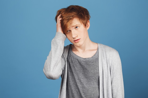 Close up portrait of unhappy young charming ginger male student in casual clothes holding hair with hand, looking aside with tired expression, having headache after getting ready for exams.