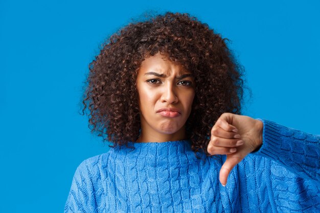 Close-up portrait unsatisfied, gloomy sad, sulking african american girl with pouting lips showing thumb-down, complaining, didnt like christmas gift, offended or insulted standing blue wall.