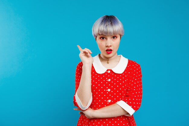 Close up portrait upset beautiful dollish girl with short light violet hair wearing red dress over blue wall