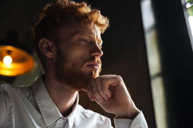 Close-up portrait of upset young redhead man holding his chin with hand, looking aside