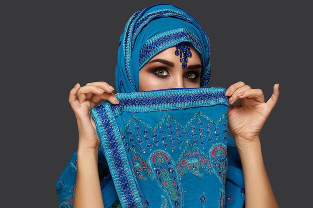 Close-up portrait of a white-skinned girl with beautiful smoky eyes wearing an elegant blue hijab decorated with sequins and jewelry. She is covering her face with a shawl and looking at the camera on