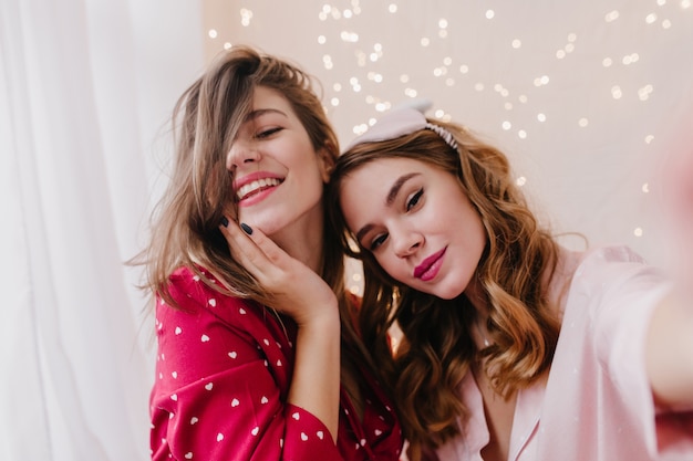 Close-up portrait of winsome curly woman making selfie with friend. Indoor photo of carefree girls taking pictures of themselves.