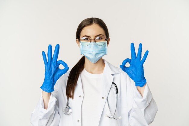 Close up portrait of woman doctor physician in face mask from coronavirus showing okay sign in appro...