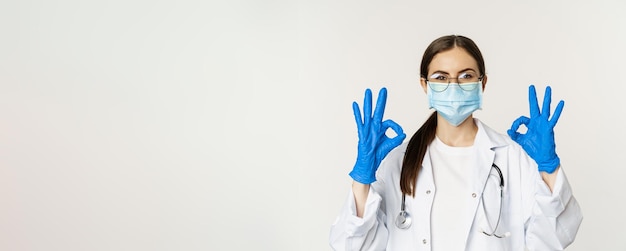 Free photo close up portrait of woman doctor physician in face mask from coronavirus showing okay sign in approval recommending praise and like smth good standing over white background