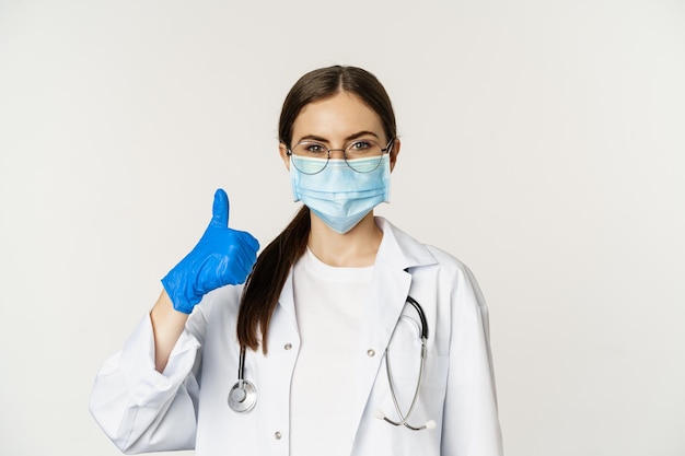 Close up portrait of woman doctor physician in face mask from coronavirus showing thumbs up and smil...