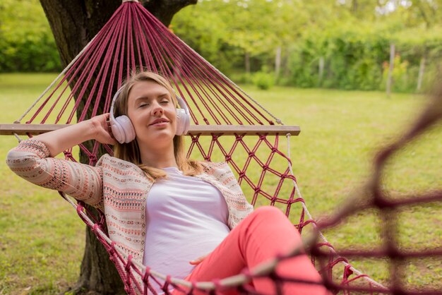 Close up portrait of woman lying down on hammock listening to music with cell phone. Cheerful girl enjoy in red hammock outdoor. woman relaxing outside listening to music with earphones