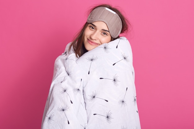 Close up portrait of woman with sleeping mask on head and wearing blanket looking smiling at camera