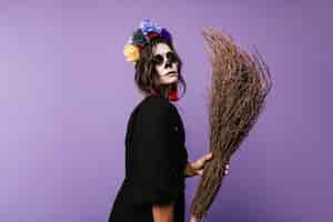 Free photo close-up portrait of woman with suspicious look in skull mask. lady in black dress holding broom.