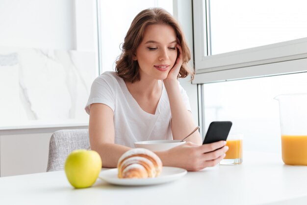 座っていると台所のテーブルで朝食をとりながら携帯電話でニュースをチェックする白いtシャツの若い笑顔の女性のクローズアップの肖像画