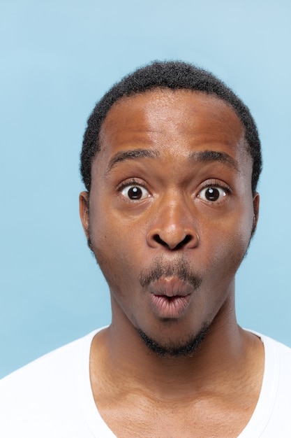 Free photo close up portrait of young african-american man in white shirt on blue wall. human emotions, facial expression, ad, sales concept.  looks shocked, astonished, wondered.