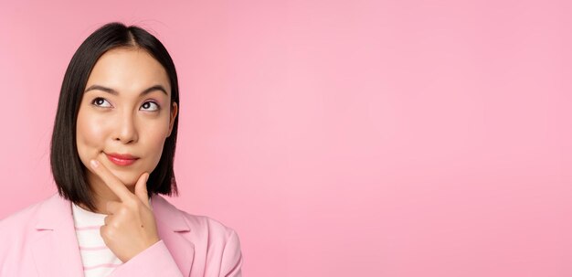 Крупный план портрета молодой азиатской бизнесвумен, думающей, задумчиво улыбающейся и смотрящей в верхний левый угол, стоящей на розовом фоне
