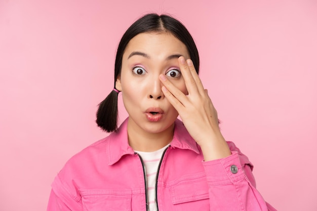 Free photo close up portrait of young asian girl looking surprised express amazement and wonder peeking through fingers standing over pink background