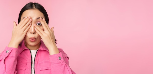 Close up portrait of young asian girl looking surprised express amazement and wonder peeking through fingers standing over pink background