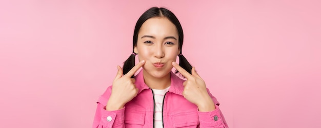 Free photo close up portrait of young asian girl showing her dimples poking cheeks silly and making funny faces standing over pink background