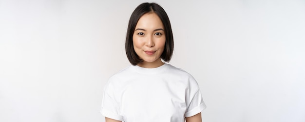 笑顔で幸せそうな白い背景を探しているTシャツを着てカメラを見て若いアジアの女性の肖像画をクローズアップ